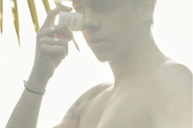 Person holding a small bottle near their eye, backlit by sunlight, with palm leaves in the background.