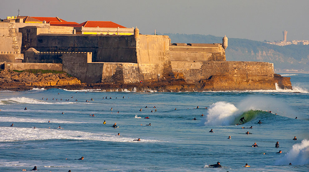 Surfers in ocean near a historic stone fort under a blue sky.
