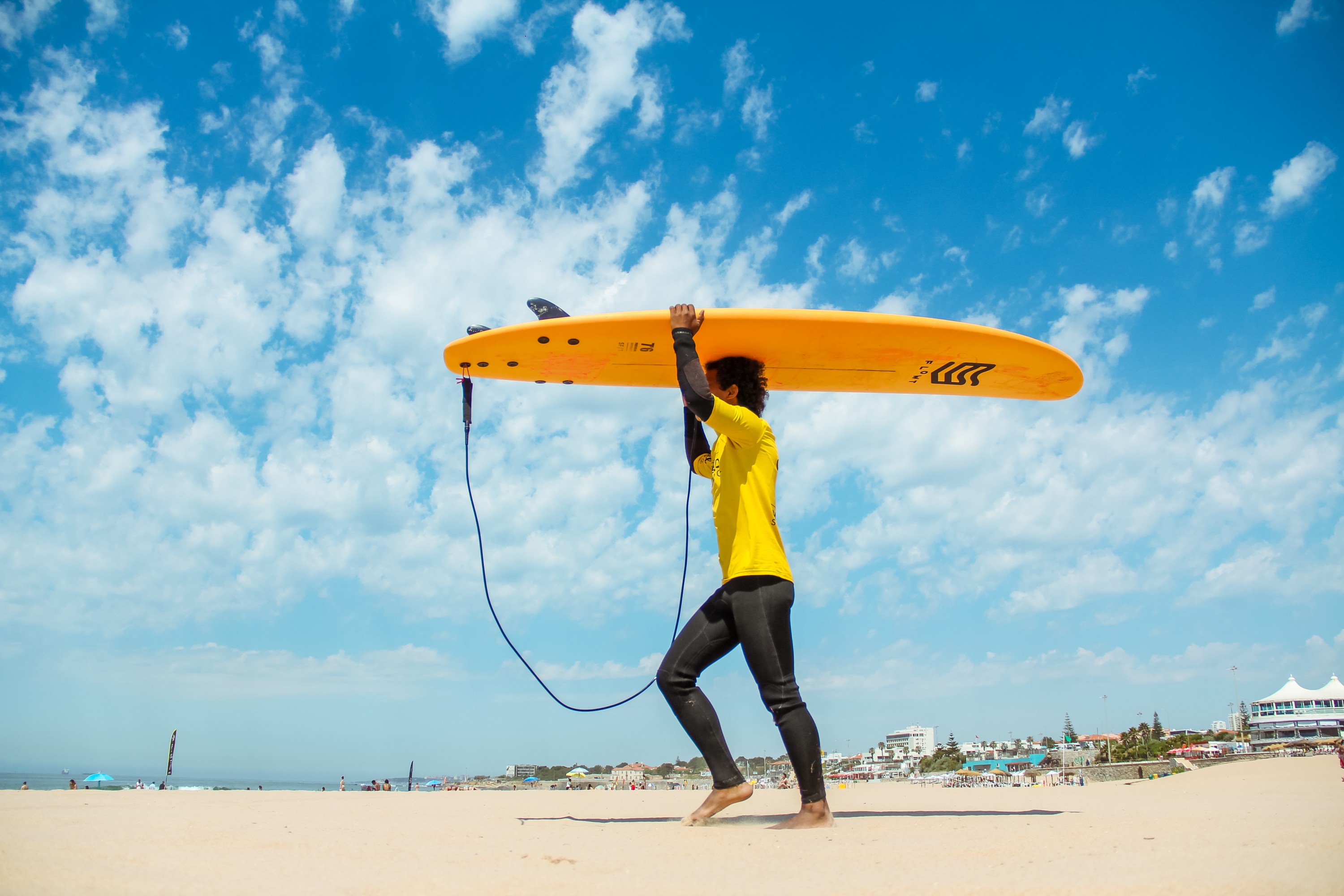 Person carrying yellow surfboard on beach under blue sky with clouds.